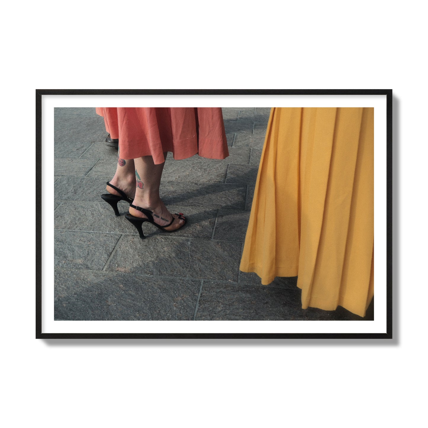 Fine art candid street photo of women in coral and mustard skirts with black heels, featuring a tattooed ankle that reads “much better”. Archival pigment print, collector size, black frame.