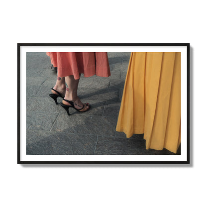 Fine art candid street photo of women in coral and mustard skirts with black heels, featuring a tattooed ankle that reads “much better”. Archival pigment print, collector size, black frame.