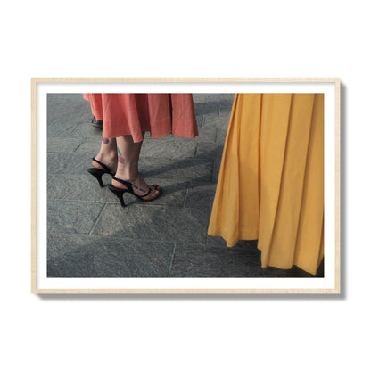 Fine art candid street photo of women in coral and mustard skirts with black heels, featuring a tattooed ankle that reads “much better”. Archival pigment print, collector size, natural wood frame.