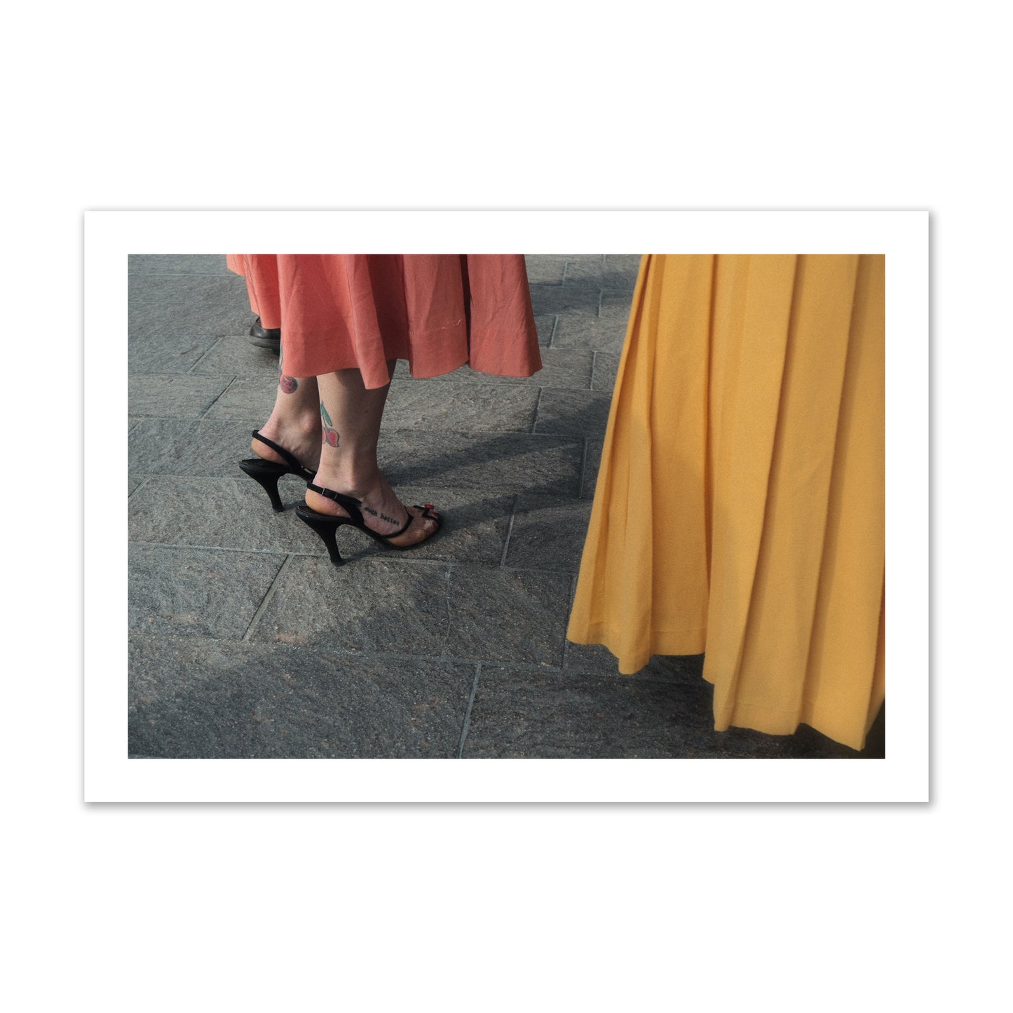 Fine art candid street photo of women in coral and mustard skirts with black heels, featuring a tattooed ankle that reads “much better”. Archival pigment print, collector size, print.