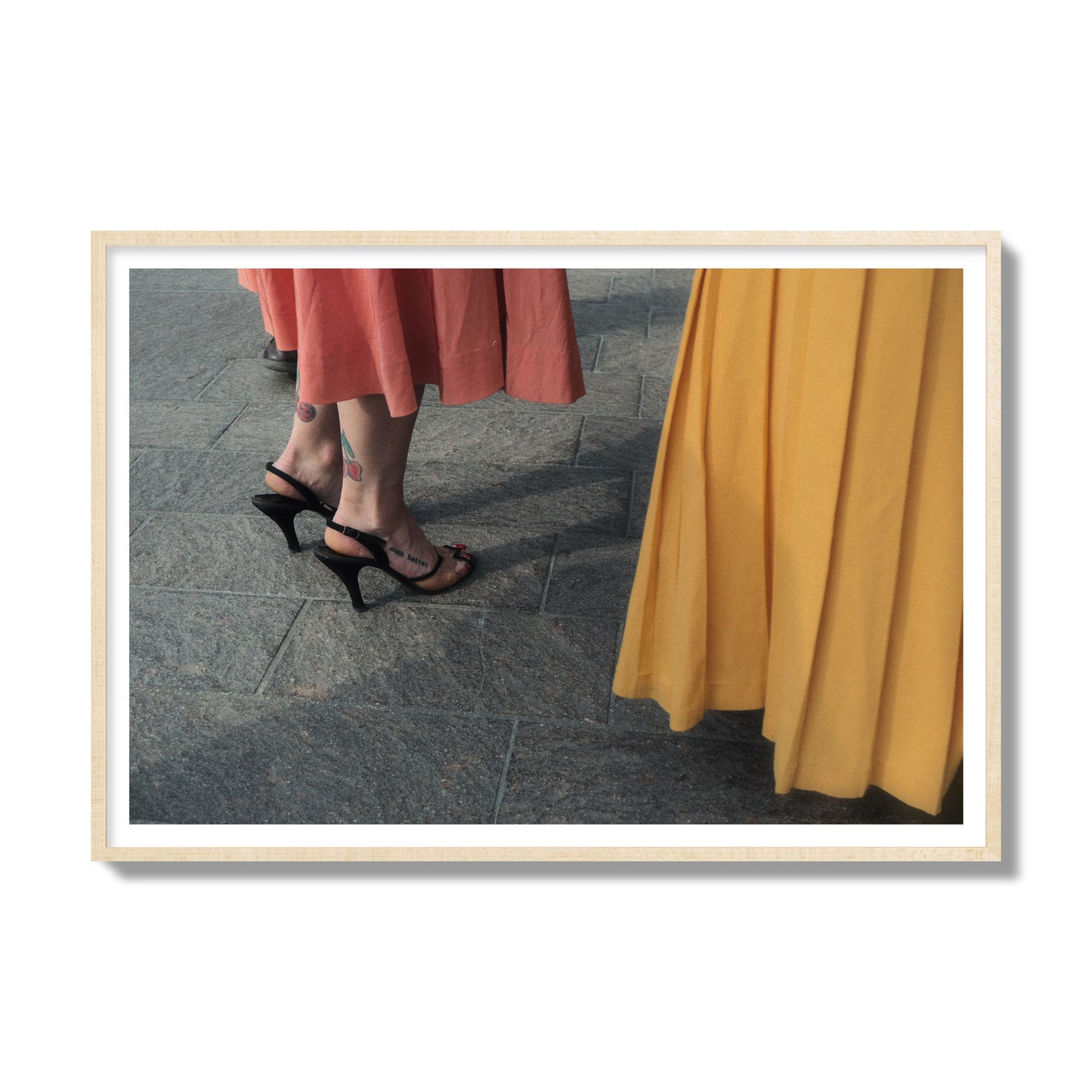 Fine art candid street photo of women in coral and mustard skirts with black heels, featuring a tattooed ankle that reads “much better”. Archival pigment print, gallery size, natural wood frame.