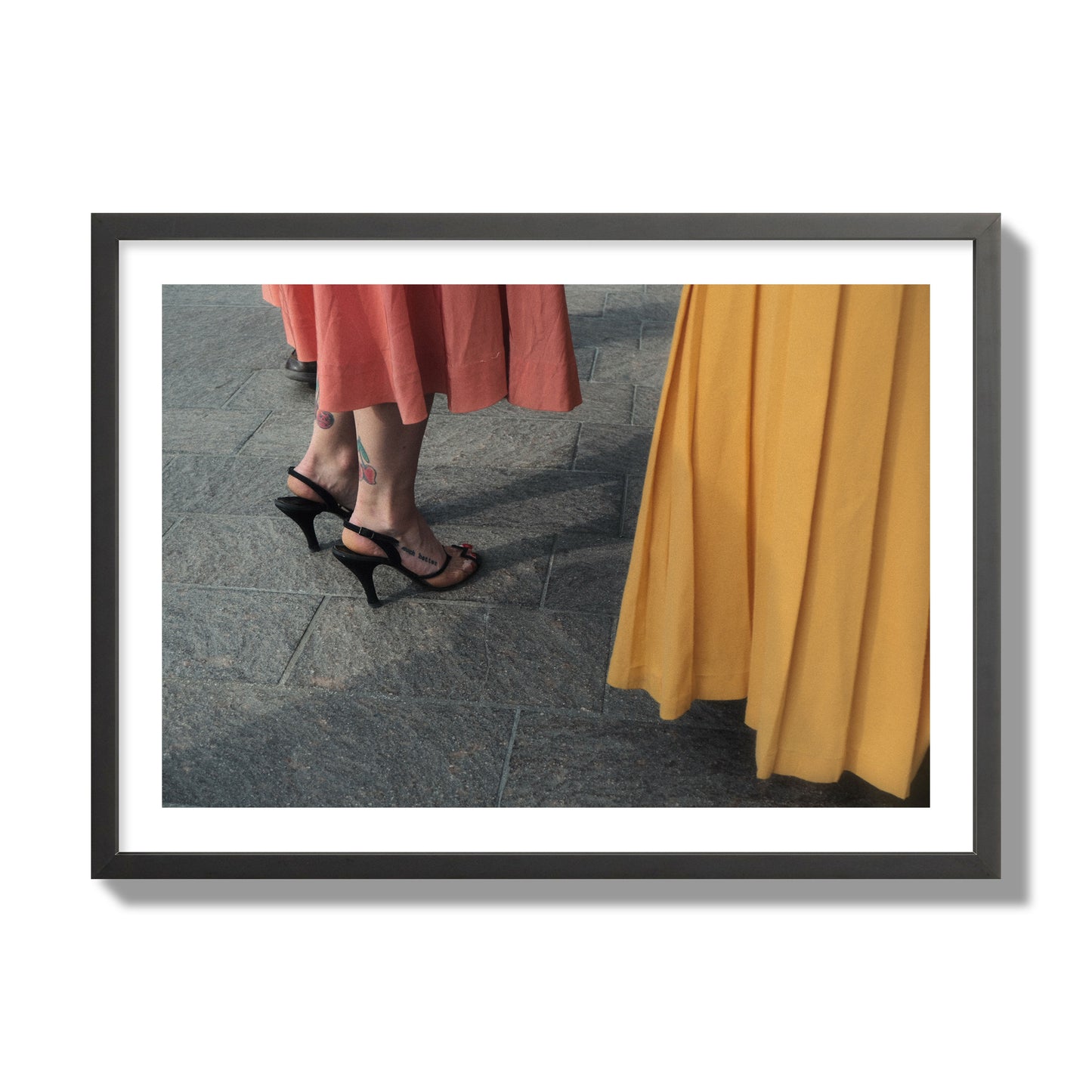 Fine art candid street photo of women in coral and mustard skirts with black heels, featuring a tattooed ankle that reads “much better”. Archival pigment print, giant size, black frame.