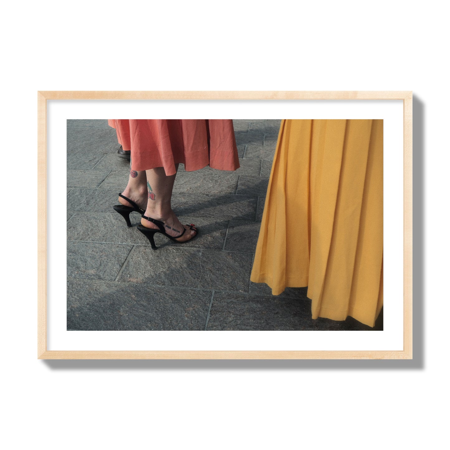 Fine art candid street photo of women in coral and mustard skirts with black heels, featuring a tattooed ankle that reads “much better”. Archival pigment print, giant size, natural wood frame.