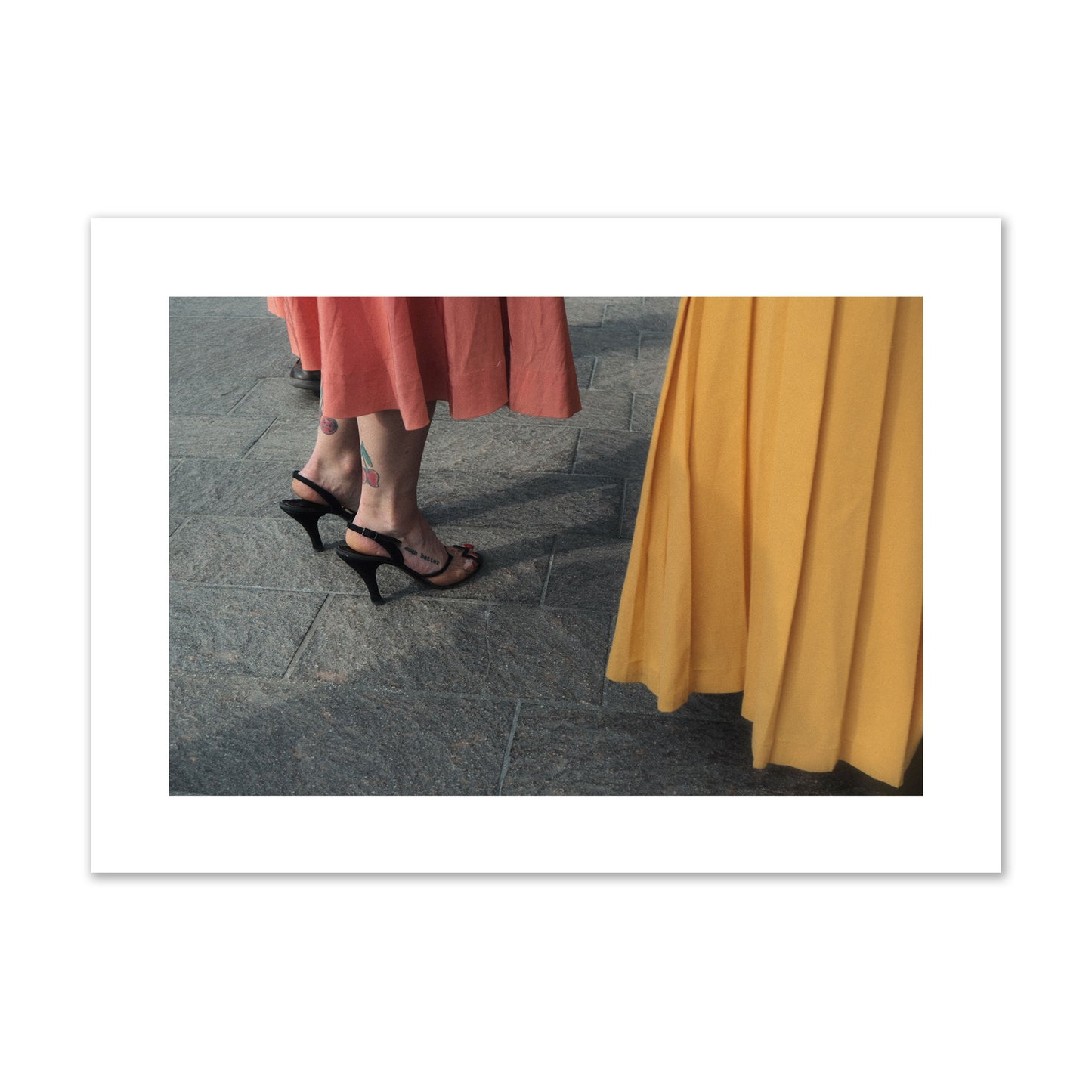 Fine art candid street photo of women in coral and mustard skirts with black heels, featuring a tattooed ankle that reads “much better”. Archival pigment print, giant size, print.
