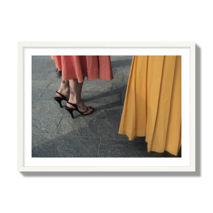 Fine art candid street photo of women in coral and mustard skirts with black heels, featuring a tattooed ankle that reads “much better”. Archival pigment print, giant size, white frame.