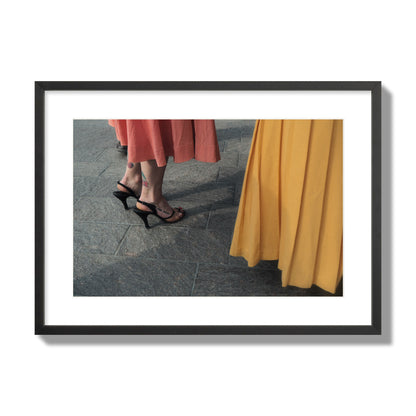 Fine art candid street photo of women in coral and mustard skirts with black heels, featuring a tattooed ankle that reads “much better”. Archival pigment print, large size, black frame.