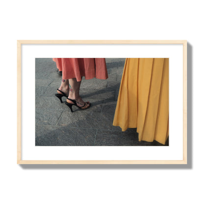 Fine art candid street photo of women in coral and mustard skirts with black heels, featuring a tattooed ankle that reads “much better”. Archival pigment print, large size, natural wood frame.
