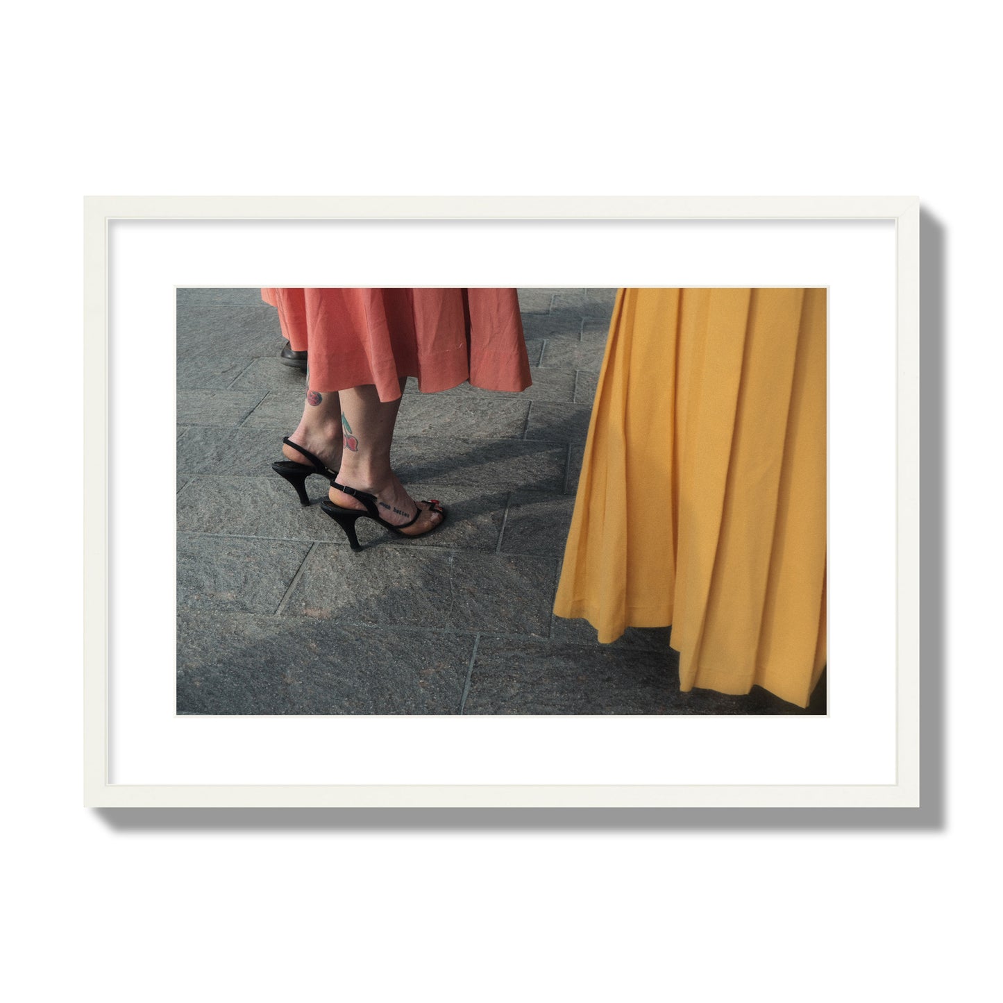 Fine art candid street photo of women in coral and mustard skirts with black heels, featuring a tattooed ankle that reads “much better”. Archival pigment print, large size, white frame.