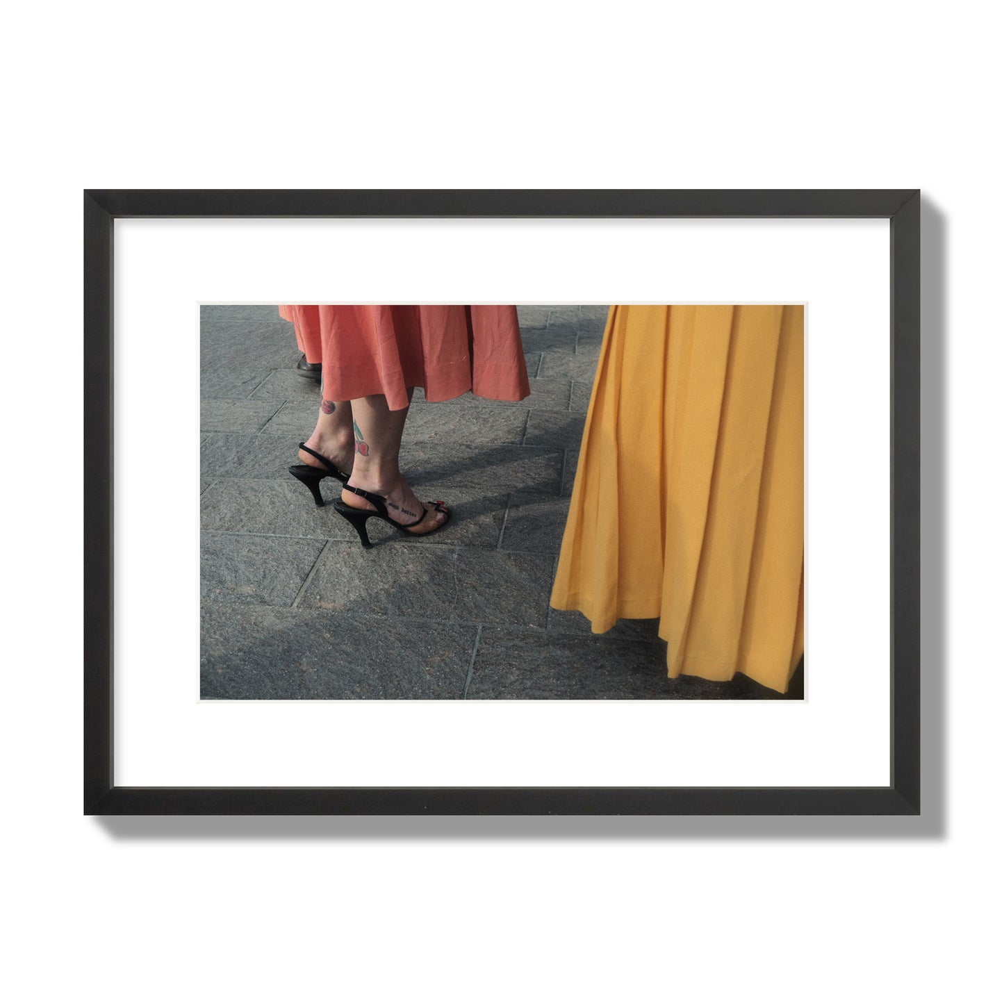 Fine art candid street photo of women in coral and mustard skirts with black heels, featuring a tattooed ankle that reads “much better”. Archival pigment print, medium size, black frame.