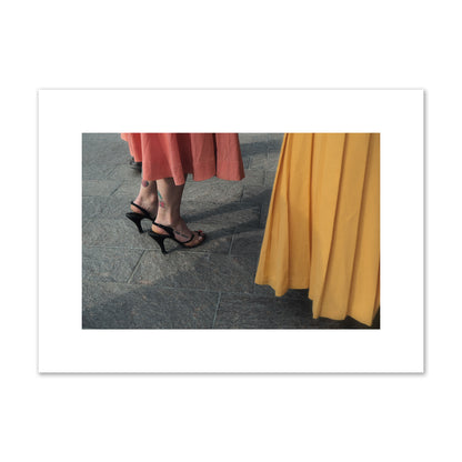 Fine art candid street photo of women in coral and mustard skirts with black heels, featuring a tattooed ankle that reads “much better”. Archival pigment print, medium size, print.