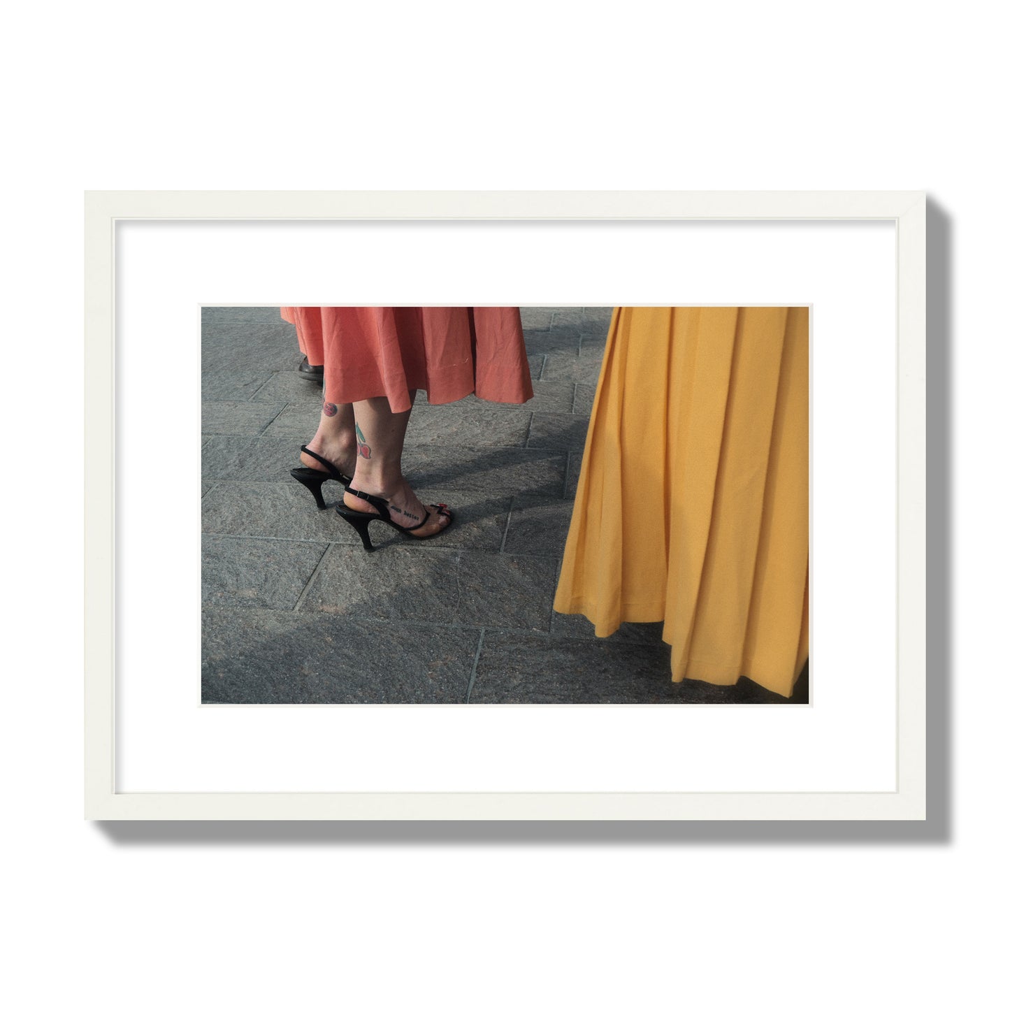 Fine art candid street photo of women in coral and mustard skirts with black heels, featuring a tattooed ankle that reads “much better”. Archival pigment print, medium size, white frame.
