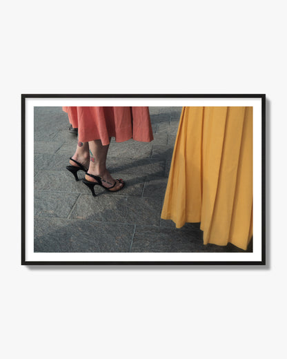 Fine art candid street photo of women in coral and mustard skirts with black heels, featuring a tattooed ankle that reads “much better”. Archival pigment print, black frame.
