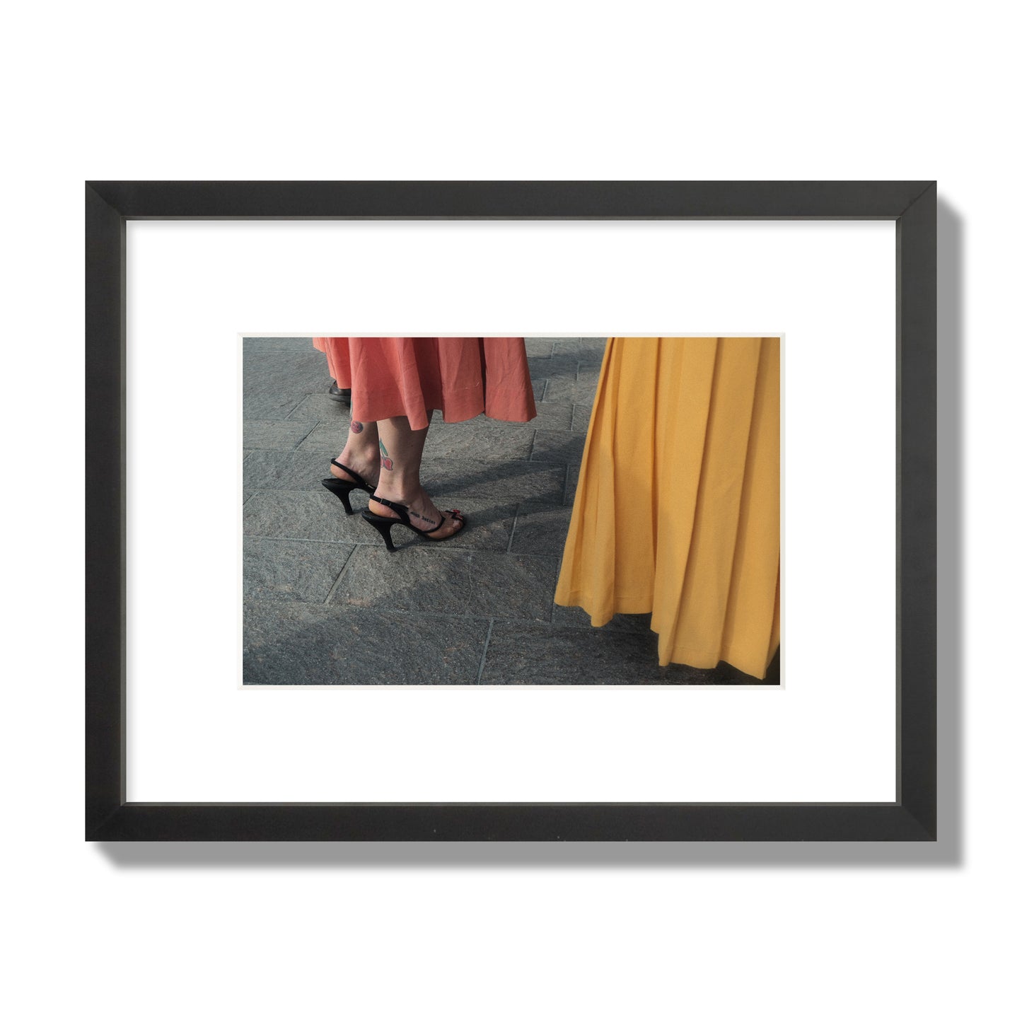 Fine art candid street photo of women in coral and mustard skirts with black heels, featuring a tattooed ankle that reads “much better”. Archival pigment print, small size, black frame.