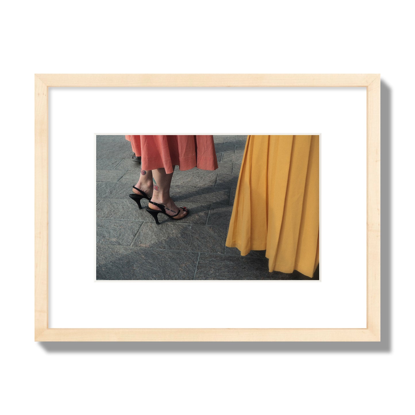 Fine art candid street photo of women in coral and mustard skirts with black heels, featuring a tattooed ankle that reads “much better”. Archival pigment print, small size, natural wood frame.