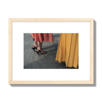 Fine art candid street photo of women in coral and mustard skirts with black heels, featuring a tattooed ankle that reads “much better”. Archival pigment print, small size, natural wood frame.