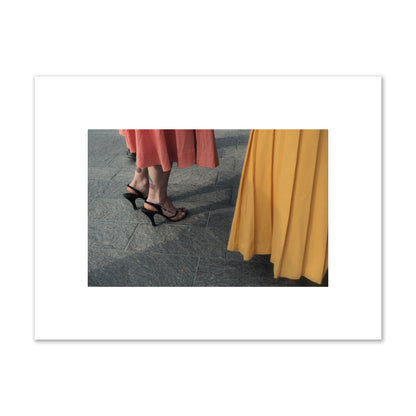 Fine art candid street photo of women in coral and mustard skirts with black heels, featuring a tattooed ankle that reads “much better”. Archival pigment print, small size, print.