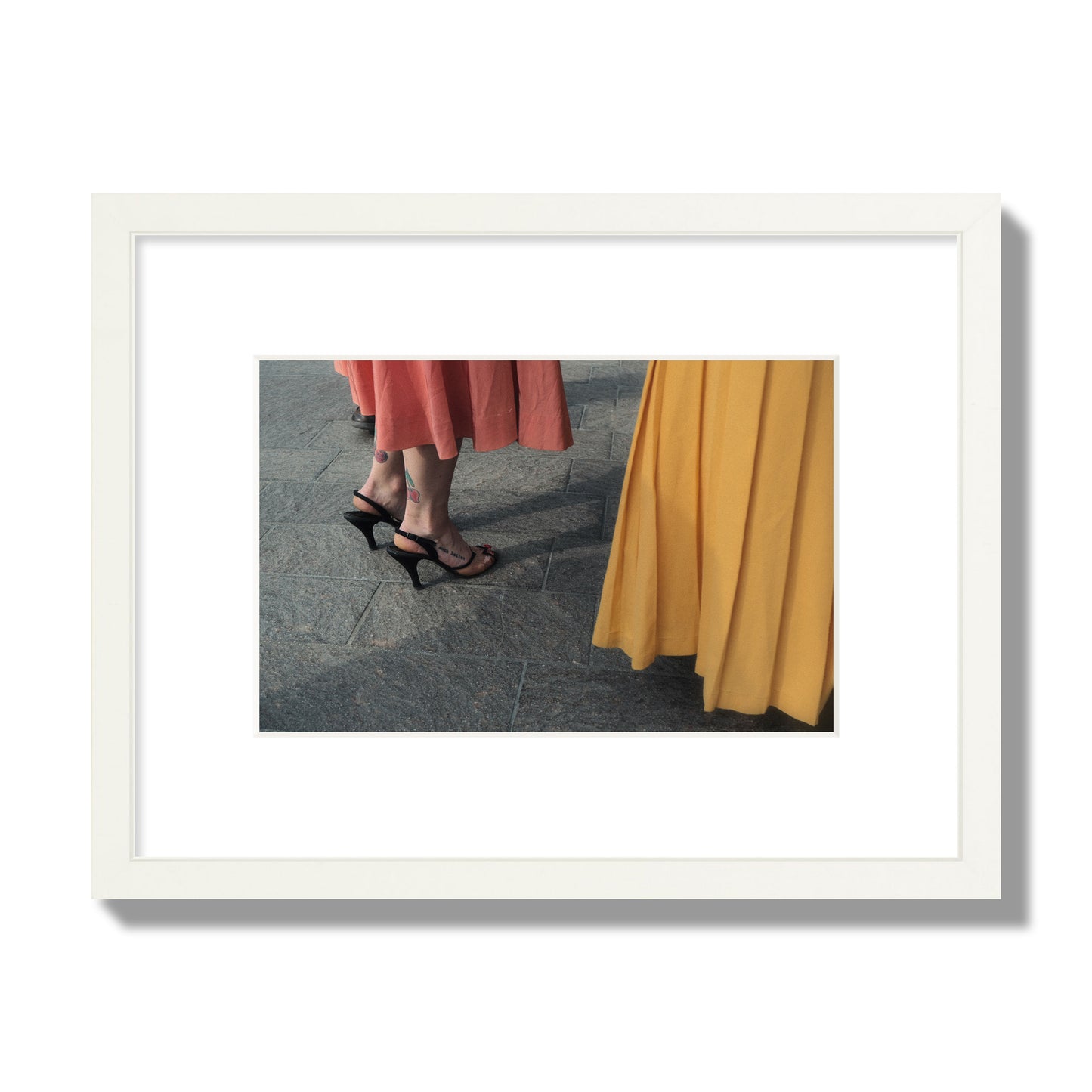 Fine art candid street photo of women in coral and mustard skirts with black heels, featuring a tattooed ankle that reads “much better”. Archival pigment print, small size, white frame.