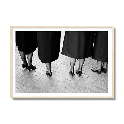 Fine art photograph depicting four women’s feet in high heels standing on a brick-paved street. One woman is notably missing a leg, adding a striking element to the composition, archival pigment print, collector size, natural wood frame.