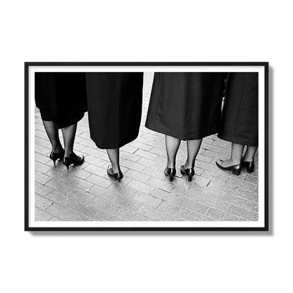 Fine art photograph depicting four women’s feet in high heels standing on a brick-paved street. One woman is notably missing a leg, adding a striking element to the composition, archival pigment print, gallery size, black frame.