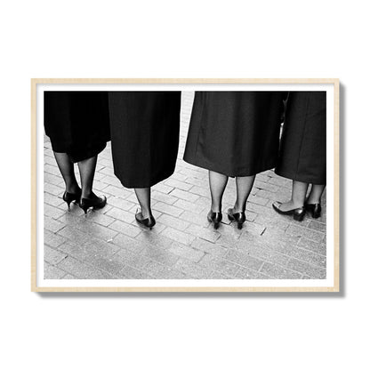 Fine art photograph depicting four women’s feet in high heels standing on a brick-paved street. One woman is notably missing a leg, adding a striking element to the composition, archival pigment print, gallery size, natural wood frame.