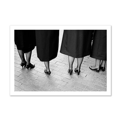 Fine art photograph depicting four women’s feet in high heels standing on a brick-paved street. One woman is notably missing a leg, adding a striking element to the composition, archival pigment print, gallery size, unframed.