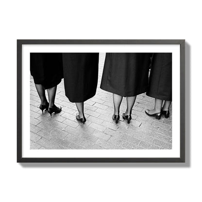 Fine art photograph depicting four women’s feet in high heels standing on a brick-paved street. One woman is notably missing a leg, adding a striking element to the composition, archival pigment print, giant size, black frame.