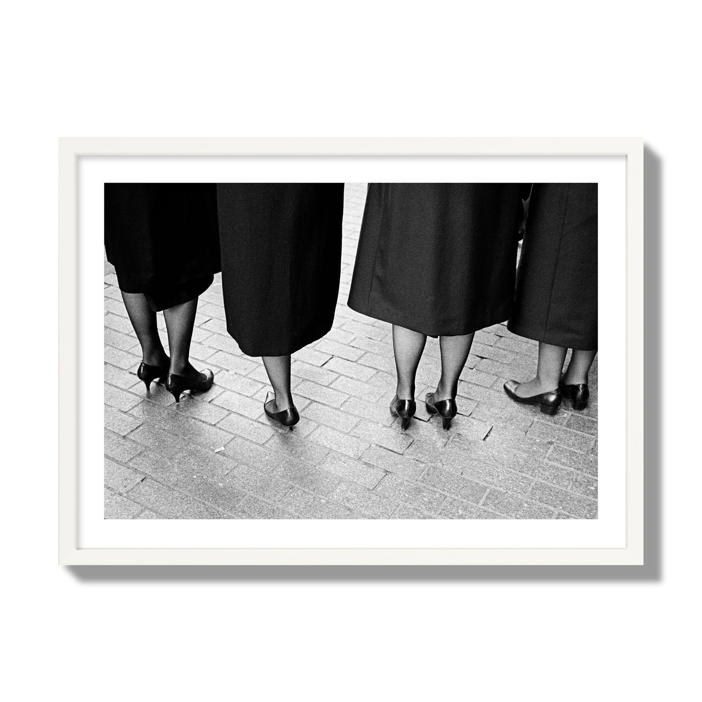 Fine art photograph depicting four women’s feet in high heels standing on a brick-paved street. One woman is notably missing a leg, adding a striking element to the composition, archival pigment print, giant size, white frame.
