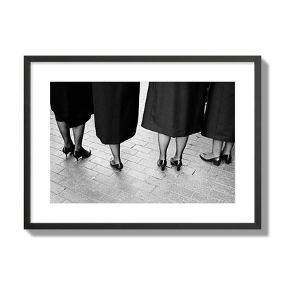 Fine art photograph depicting four women’s feet in high heels standing on a brick-paved street. One woman is notably missing a leg, adding a striking element to the composition, archival pigment print, large size, black frame