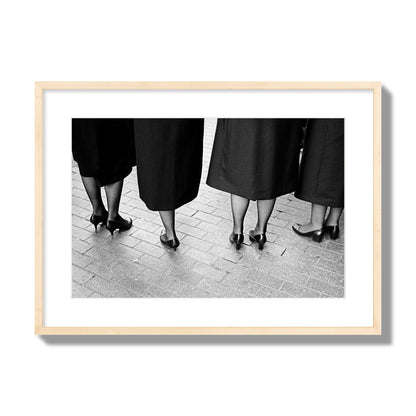 Fine art photograph depicting four women’s feet in high heels standing on a brick-paved street. One woman is notably missing a leg, adding a striking element to the composition, archival pigment print, large size, natural wood frame