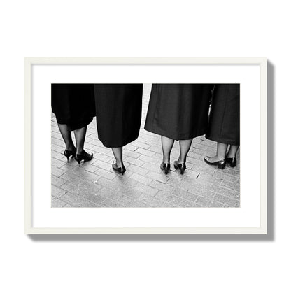 Fine art photograph depicting four women’s feet in high heels standing on a brick-paved street. One woman is notably missing a leg, adding a striking element to the composition, archival pigment print, large size, white frame