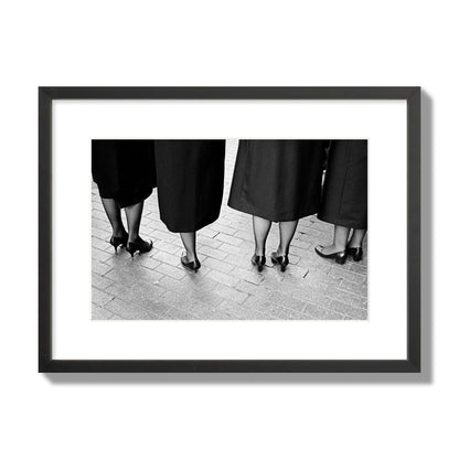 Fine art photograph depicting four women’s feet in high heels standing on a brick-paved street. One woman is notably missing a leg, adding a striking element to the composition, archival pigment print, medium size, black frame