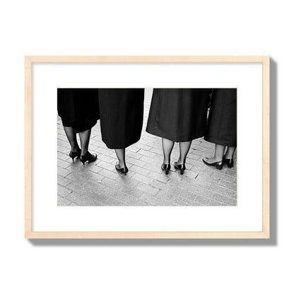 Fine art photograph depicting four women’s feet in high heels standing on a brick-paved street. One woman is notably missing a leg, adding a striking element to the composition, archival pigment print, medium size, natural wood frame