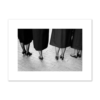 Fine art photograph depicting four women’s feet in high heels standing on a brick-paved street. One woman is notably missing a leg, adding a striking element to the composition, archival pigment print, medium size, unframed.