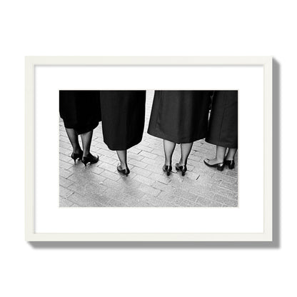 Fine art photograph depicting four women’s feet in high heels standing on a brick-paved street. One woman is notably missing a leg, adding a striking element to the composition, archival pigment print, medium size, white frame
