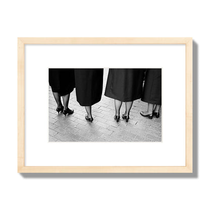 Fine art photograph depicting four women’s feet in high heels standing on a brick-paved street. One woman is notably missing a leg, adding a striking element to the composition, archival pigment print, small size, natural wood frame