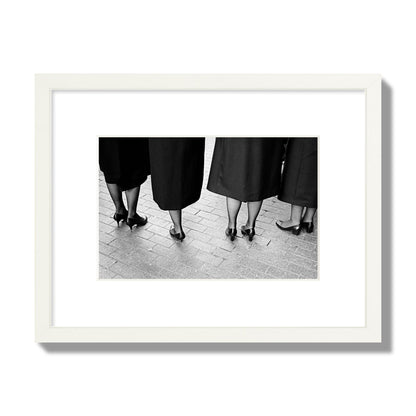 Fine art photograph depicting four women’s feet in high heels standing on a brick-paved street. One woman is notably missing a leg, adding a striking element to the composition, archival pigment print, small size, white frame