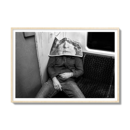 Fine art photograph of a man asleep on the subway with a newspaper covering their face, archival pigment print, gallery size, natural wood frame.