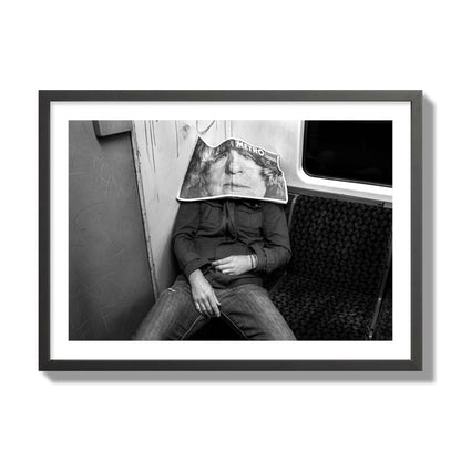 Fine art photograph of a man asleep on the subway with a newspaper covering their face, archival pigment print, giant size, black frame.