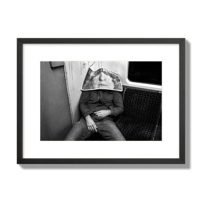 Fine art photograph of a man asleep on the subway with a newspaper covering their face, archival pigment print, medium size, black frame.