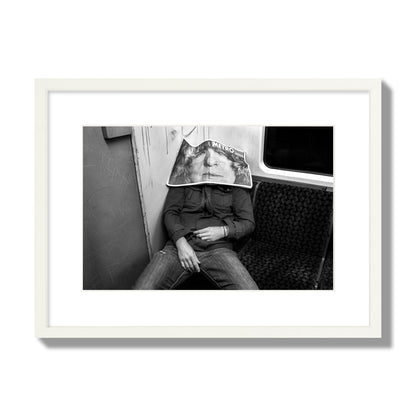 Fine art photograph of a man asleep on the subway with a newspaper covering their face, archival pigment print, medium size, white frame.