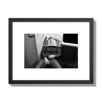 Fine art photograph of a man asleep on the subway with a newspaper covering their face, archival pigment print, small size, black frame.