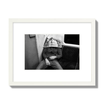 Fine art photograph of a man asleep on the subway with a newspaper covering their face, archival pigment print, small size, white frame.