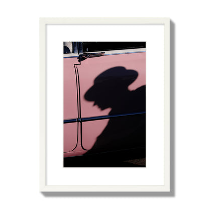 Fine art photograph of a man's shadow cast on a pink retro car, archival pigment print, medium size, white frame.