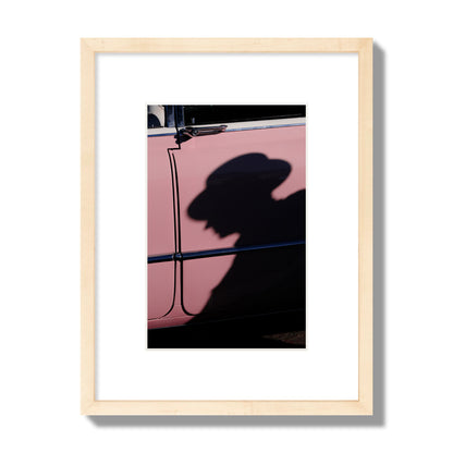 Fine art photograph of a man's shadow cast on a pink retro car, archival pigment print, small size, natural wood frame.