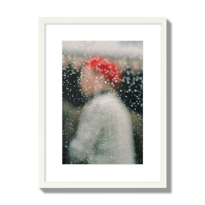 Fine art photograph of a woman with a red hat through raindrops on a window, archival pigment print, medium size, white frame.