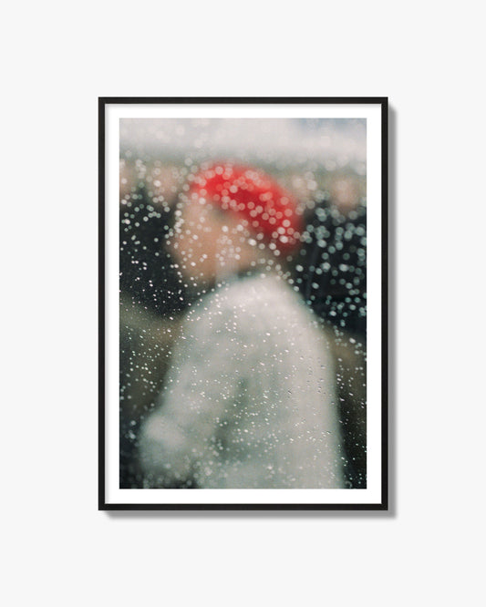 Fine art photograph of a woman with a red hat through raindrops on a window, archival pigment print, black frame.