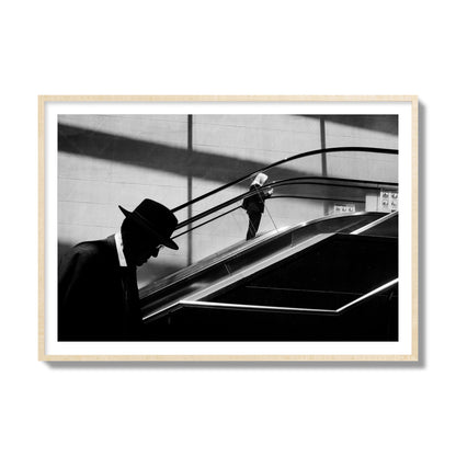 Fine art black and white street photograph of a man ascending an escalator. Archival pigment print, collector size, natural wood frame.