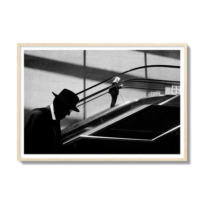 Fine art black and white street photograph of a man ascending an escalator. Archival pigment print, gallery size, natural wood frame.