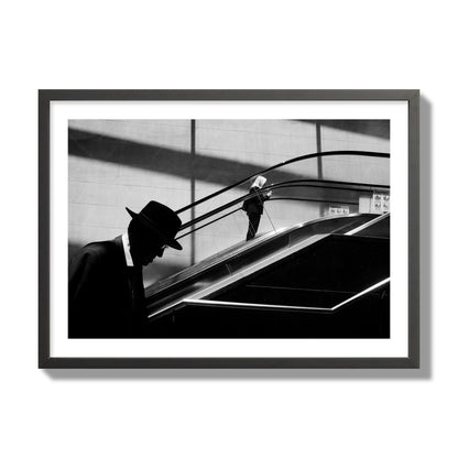 Fine art black and white street photograph of a man ascending an escalator. Archival pigment print, giant size, black frame.