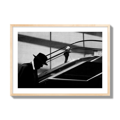 Fine art black and white street photograph of a man ascending an escalator. Archival pigment print, giant size, natural wood frame.